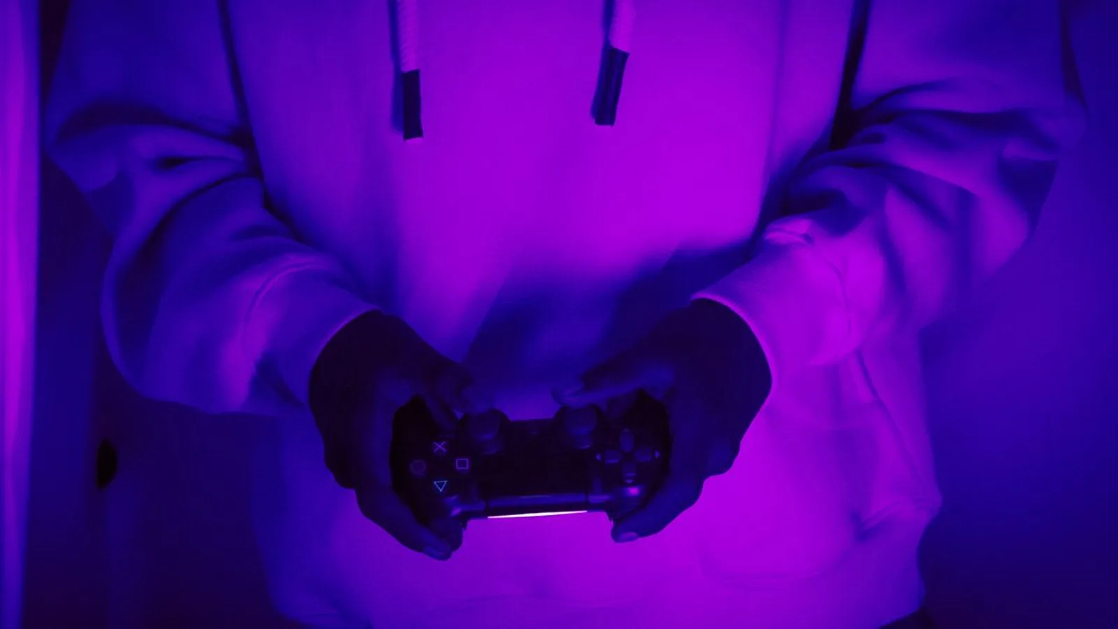 A person in a hoodie holds a game controller under neon purple light.