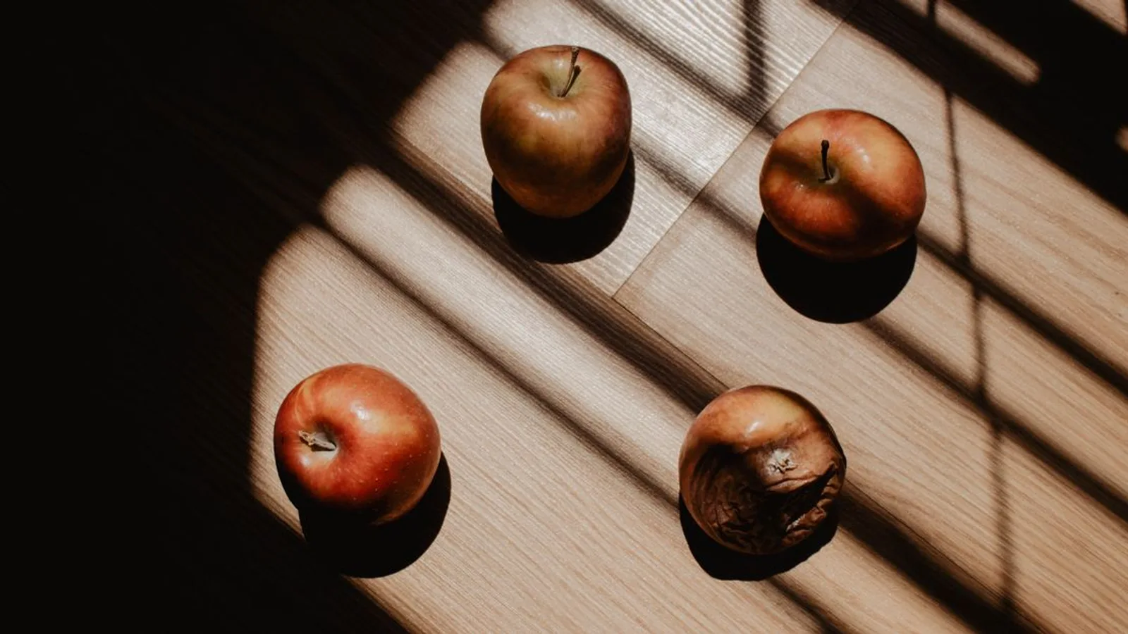 A striking artistic still life featuring three fresh red apples and one rotten apple on a wooden surface with dramatic shadows.