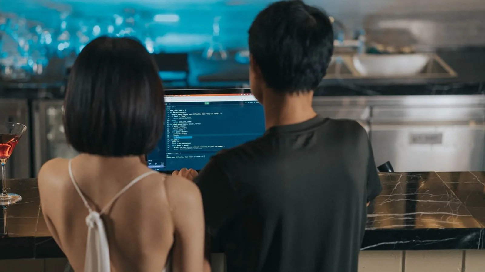 Couple coding together at a bar counter, drinks nearby, casual atmosphere.